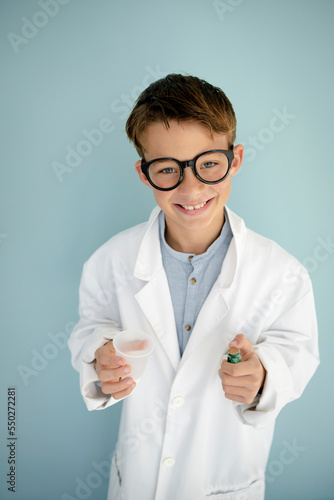 young cool boy ,with big massive black glasses is dressed up as a mad scientist holding beaker and vials in hand in front of blue background