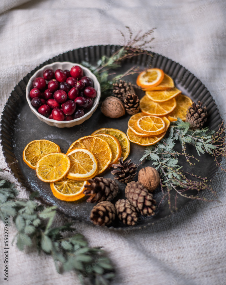 Weihnachtsdeko auf einem Teller aus Metall, eine Schale mit Cranberrys, getrocknete Orangenscheiben, Zapfen, Nüsse und Mimosenzweig, dekorieren im Advent