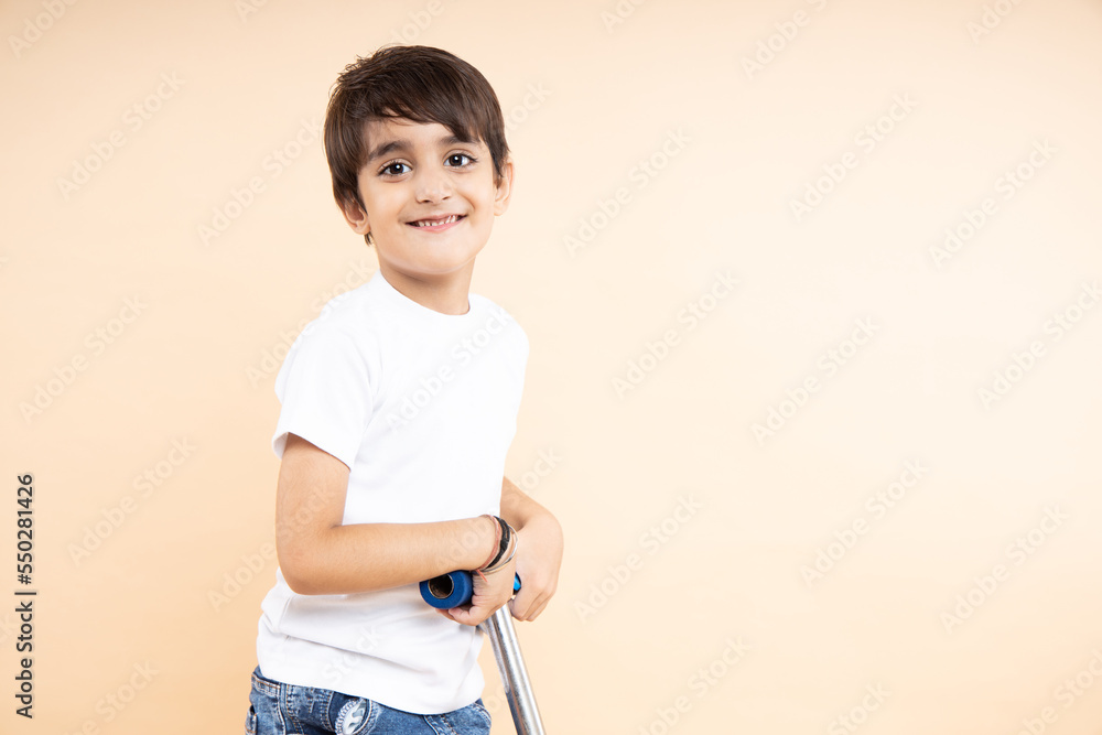 Closeup of smiling indian boy kid wearing white t-shirt rides holding scooter or bicycle handle on beige background with copy space. Studio shot, Childhood and active leisure concept. Closeup of smiling indian boy kid wearing white t-shirt rides holding scooter or bicycle handle on beige background with copy space. Studio shot, Childhood and active leisure concept.