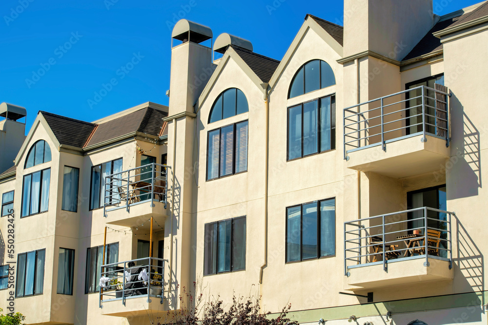 Beige house or home facade with visible windows and balconies and chimney vents with brown roof tiles and front yard trees