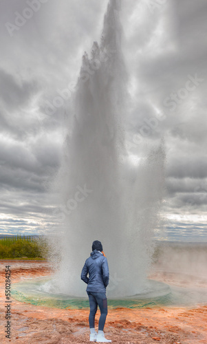 A young woman in sporty clothes watches Eruption of Strokkur Geyser in Haukadalur Valley - Iceland