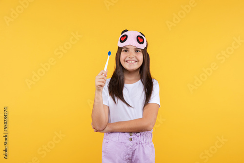  Studio shot of smiling little girl in white t-shirt and eye mask holding  toothbrush looking to the camera. Morning routin and kids hygiene concept.