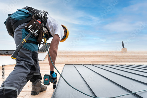 A roofer with a safety harness and tool belt is working with a electric screwdriver on the roof. He is anchoring the metal roofing with a screws. photo