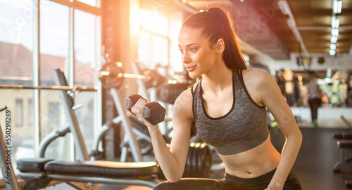 Young active sporty woman flexing muscles with dumbbell while sitting on weight bench in the gym.
