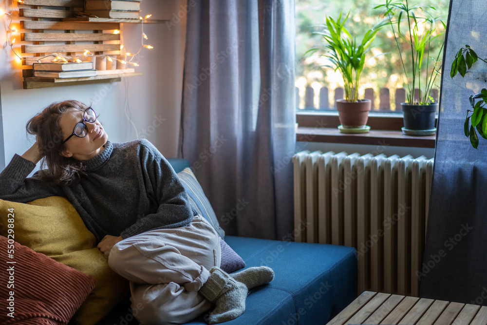 Young adult happy cheerful woman 30s in warm sweater and eyeglasses sitting on sofa enjoying resting and relaxing with her eyes closed