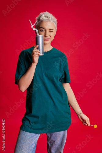 Portrait of young stylish woman with short hair posing in comfortabel casual clothes, drinking coke isolated over red background photo