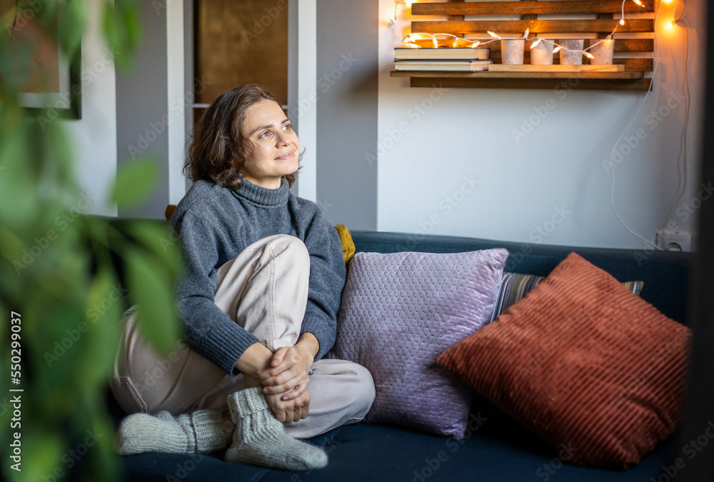 Young adult happy cheerful woman in warm sweater sitting on sofa enjoying resting and relaxing