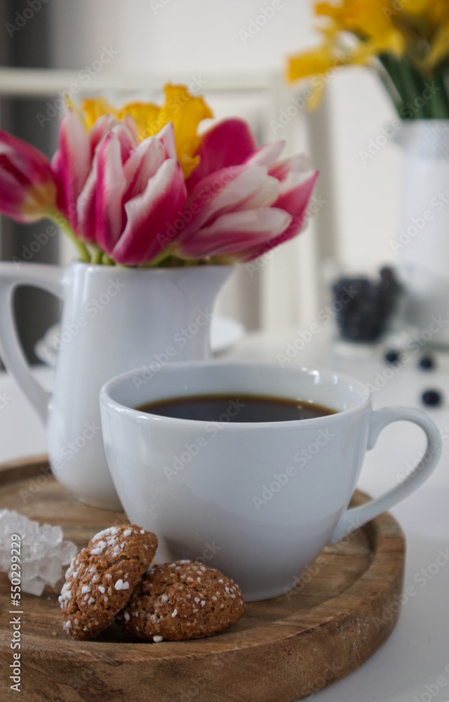A cup of coffee and a vase with tulips and daffodils on a wooden stand on a table in the kitchen A cup of coffee and a vase with tulips and daffodils on a wooden stand on a table in the kitchen