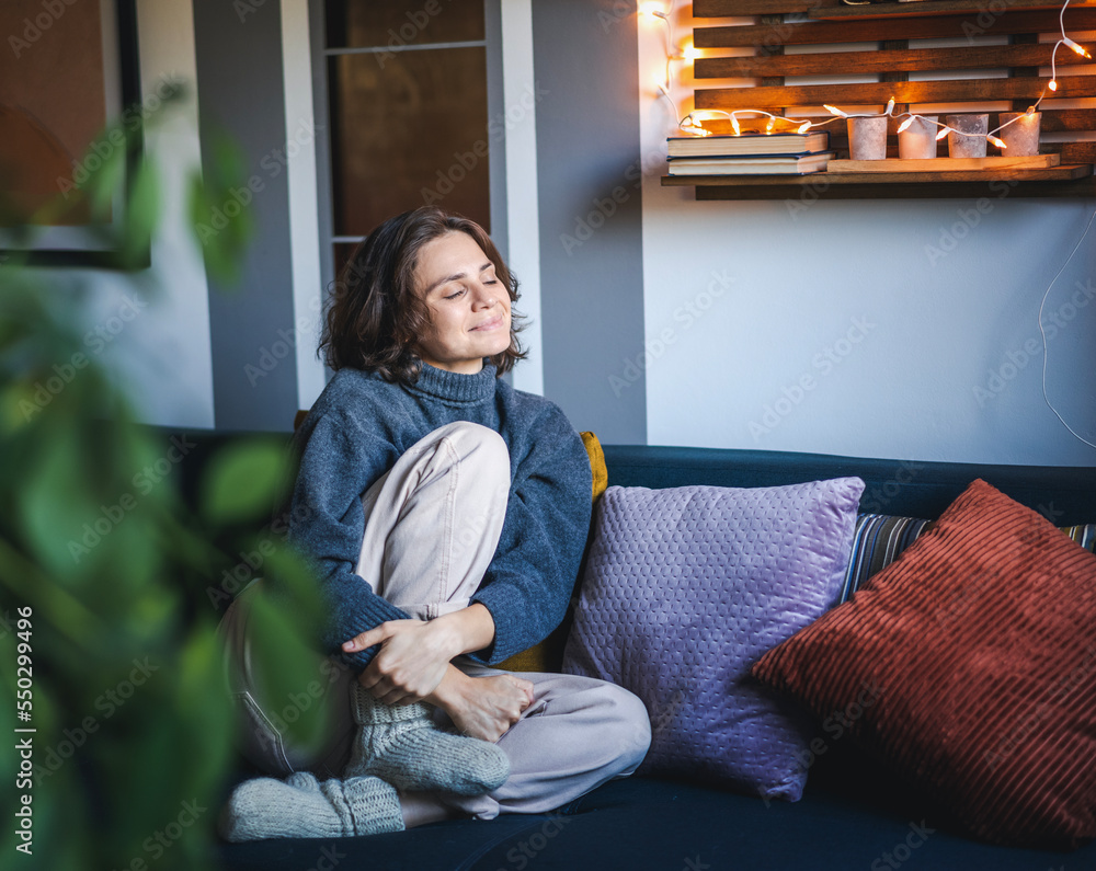 Young adult happy cheerful woman in warm sweater sitting on sofa enjoying resting and relaxing with her eyes closed
