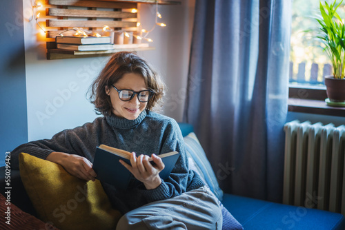 Obraz na płótnie Young smiling cheerful woman in a warm sweater and eyeglasses reading a book while sitting on the couch in the room w salonie