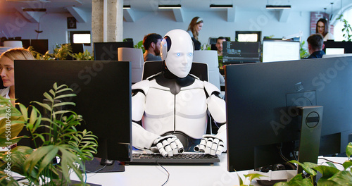 White robot sitting at table in office and typing on keyboard. Artificial intelligence working at computer in coworking space. Humanoid IT worker. Call center. Futuristic employee texting. Technology.