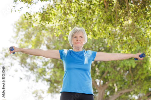 Donna anziana con i capelli grigi e un maglietta sportiva celeste fa esercizio fisico con dei pesi in un parco.