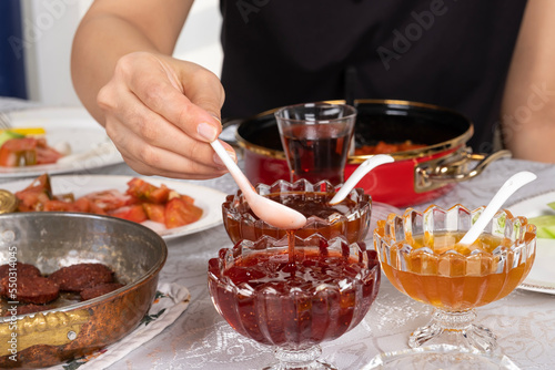 Strawberry jam, caucasian woman hand holding spoon to take some strawberry jam. Traditional, delicious rich Turkish breakfast table. Tea in glass, sausage called sucuk in fry pan Tomatoes, cheese.