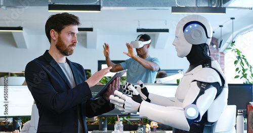 Man programming robot with tablet computer in hands in office. Male developer checking work of android by device. Humanoid moving and executing instructions and orders. Artificial intelligence startup