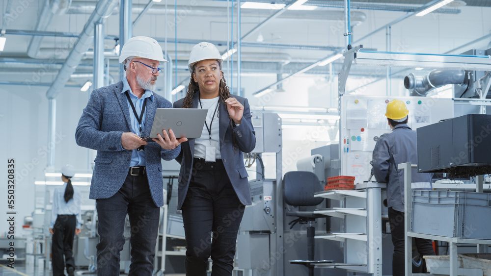 Two Heavy Industry Engineers in Hard Hats Walking with Laptop Computer and Talking in a Factory. Footage of Two Manufacturing Employees at Work in Production Facility. Two Heavy Industry Engineers in Hard Hats Walking with Laptop Computer and Talking in a Factory. Footage of Two Manufacturing Employees at Work in Production Facility.
