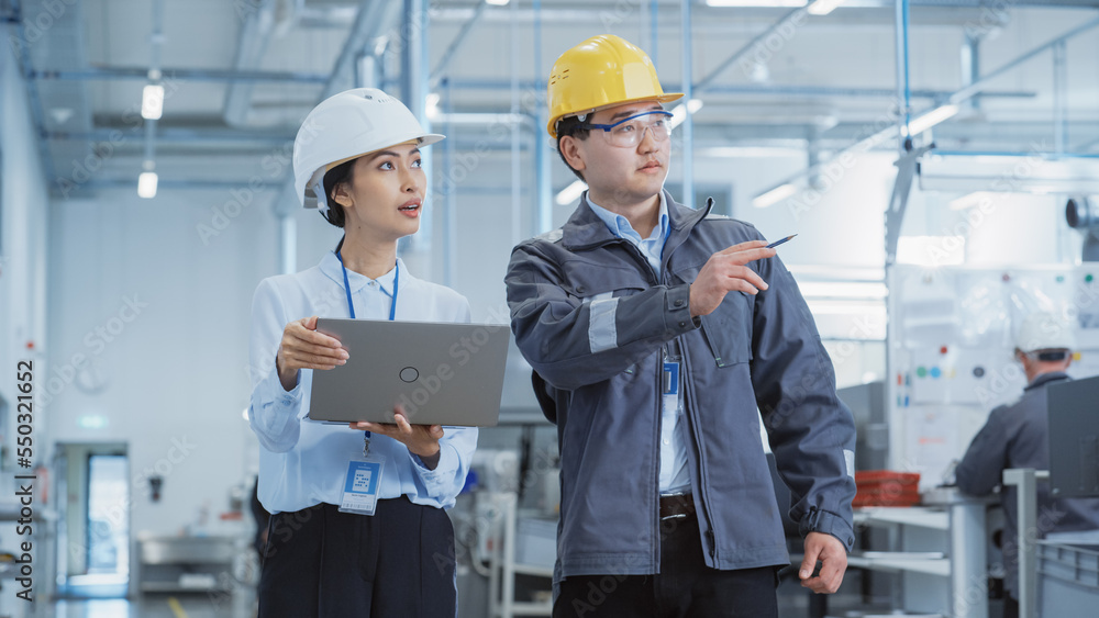 Two Young Asian Heavy Industry Engineers in Hard Hats Walking with Laptop Computer and Talking in a Factory. Two Manufacturing Employees at Work in Research and Development Facility. Two Young Asian Heavy Industry Engineers in Hard Hats Walking with Laptop Computer and Talking in a Factory. Two Manufacturing Employees at Work in Research and Development Facility.