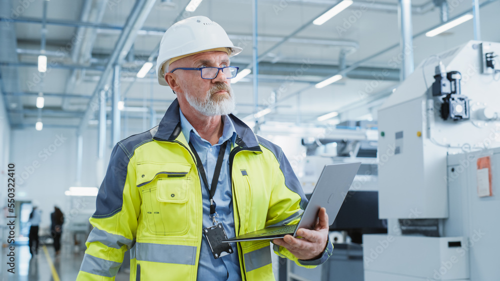 Portrait of a Bearded Middle Aged Engineer Standing in a Factory Facility, Wearing a High Visibility Jacket and a White Hard Hat. Heavy Industry Specialist Working on Laptop Computer.