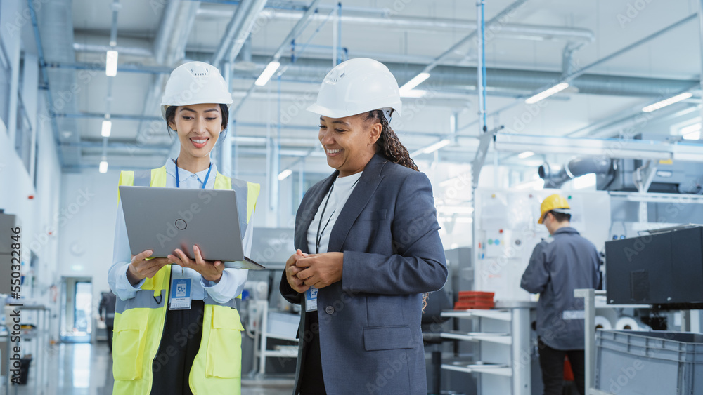 Portrait of Two Female Employees in Hard Hats at Factory Discussing Assignments at Industrial Machine Facility, Using Laptop Computer. Smiling Asian Engineer and African American Technician at Work. Portrait of Two Female Employees in Hard Hats at Factory Discussing Assignments at Industrial Machine Facility, Using Laptop Computer. Smiling Asian Engineer and African American Technician at Work.