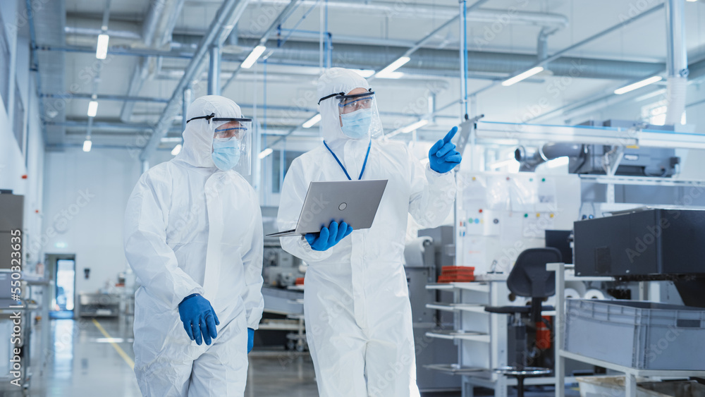 Two Scientists Walking in a Heavy Industry Factory in Sterile Coveralls and Face Masks, Using Laptop Computer. Examining Industrial Machine Settings and Configuring Production Functionality. Two Scientists Walking in a Heavy Industry Factory in Sterile Coveralls and Face Masks, Using Laptop Computer. Examining Industrial Machine Settings and Configuring Production Functionality.