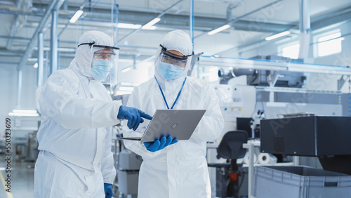 Foto Two Scientists Walking in a Heavy Industry Factory in Sterile Coveralls and Face Masks, Using Laptop Computer