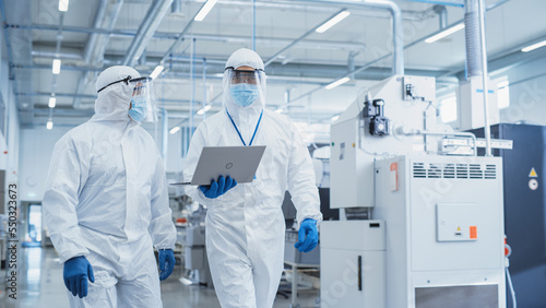 Εκτύπωση καμβά Two Scientists Walking in a Heavy Industry Factory in Sterile Coveralls and Face Masks, Using Laptop Computer