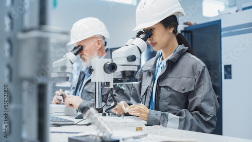 Electronic Manufacturing Factory: Two Engineers Examining Small Scale Industrial Equipment Parts Through a Microscope. Technicians Testing New Factory Components for Production Quality.