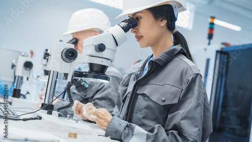 Young Asian Industrial Scientist and Older Engineer Working at a Desk in a Factory Facility, Using Microscopes to Inspect the Manufacturing Production Parts for Quality.