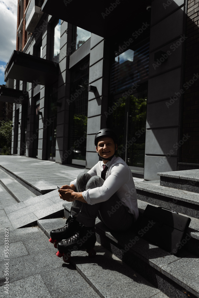 happy businessman in roller skates and helmet holding smartphone and smiling at camera while sitting on stairs near building.