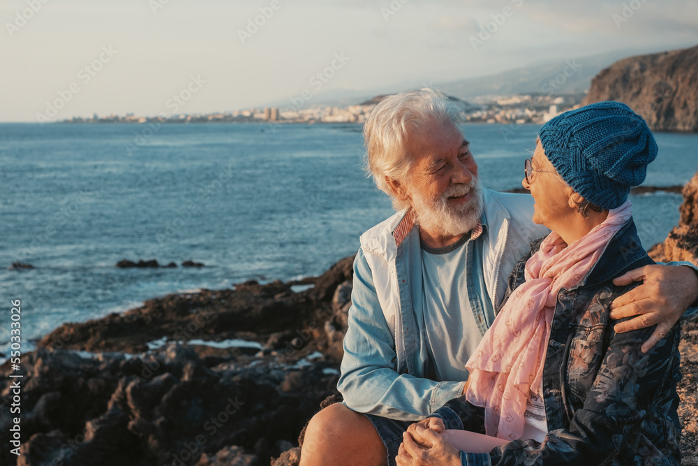 Smiling beautiful senior couple sitting on the rocks at sea enjoying travel and nature at sunset light. Relaxed lifestyle for a caucasian couple of retirees