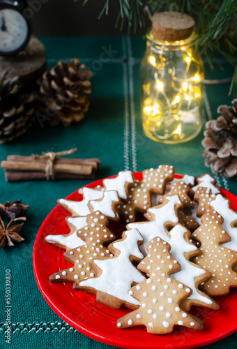 Festive cookies decorated with white icing on a red plate on a green background with lights. Homemade ginger and cinnamon Christmas cookies. Vertical orientation. Selective focus.