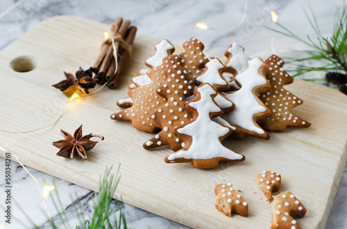 Festive cookies decorated with white icing on a wooden cutting board on a marble background with lights. Homemade ginger and cinnamon Christmas cookies. Horizontal orientation.