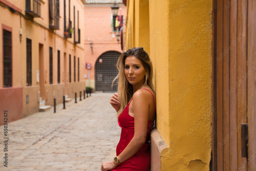 Young, blonde, beautiful woman in a red dress is visiting seville. The woman poses for the camera very elegant and like a model in the typical streets of the city. Holidays and travels