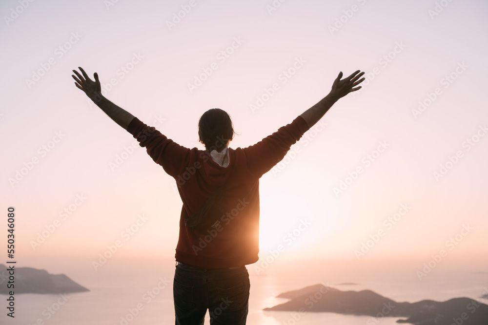 Fototapeta premium A young man meets the sunset, arms outstretched in a free gesture on a mountain by the sea.