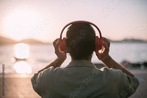 Portrait from the back of a young man in bright big headphones by the sea at sunset. A handsome guy listens to music on the ocean in the rays of the sun, with his back to the camera