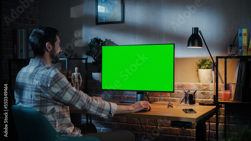 Young Attractive Man Working from Home on Desktop Computer with Green Screen Mock Up Display. Male Checking Corporate Accounts, Messaging Colleagues. Loft Living Room in the Evening