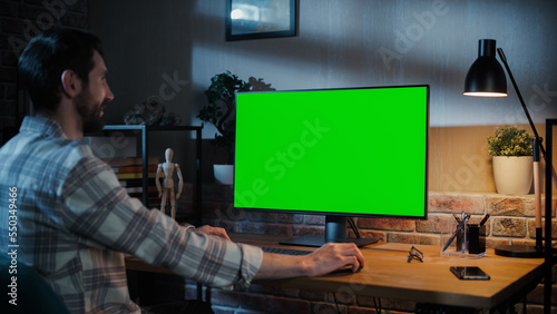 Young Handsome Man Working from Home on Desktop Computer with Green Screen Mock Up Display. Creative Male Checking Social Media, Browsing Internet. Loft Living Room in the Evening