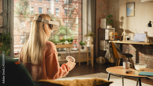 Female with Blond Hair Using Virtual Reality Headset and Controllers at Home. Creative Woman Sitting on a Sofa in Living Room, Playing VR Video Game or Working in Special Metaverse Software.