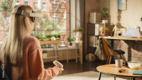 Female with Blond Hair Using Virtual Reality Headset and Controllers at Home. Creative Woman Sitting on a Couch in Living Room, Playing VR Video Game or Working on a Work Task.