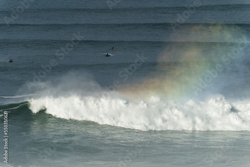 Big giant waves crashing in Nazare, Portugal creating a rainbow in the ocean. Surfers and jet skis in the water. Biggest waves in the world. Touristic destination for surfing.