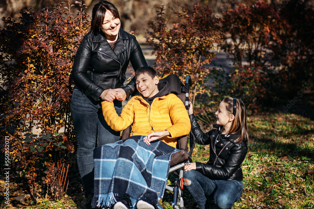 Diversity and inclusion. Happy family, mother, daughter and son teen boy with cerebral palsy spending time together in autumn fall park. Teen boy who uses a wheelchair walking with family - obrazy, fototapety, plakaty 