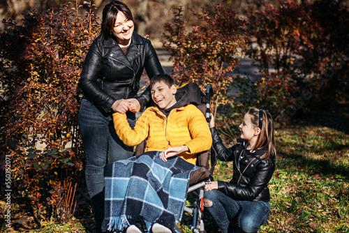 Diversity and inclusion. Happy family, mother, daughter and son teen boy with cerebral palsy spending time together in autumn fall park. Teen boy who uses a wheelchair walking with family photo