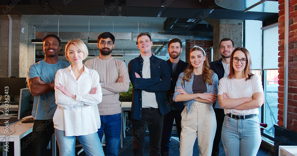 Portrait shot of young successful diverse people standing in office, smiling to camera and crossing hands. Multiethnic men and women. Group of IT startup and communication, management strategy.