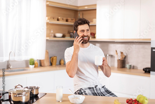 Happy adult caucasian guy with beard in white t-shirt drinks cup of coffee, calls by phone