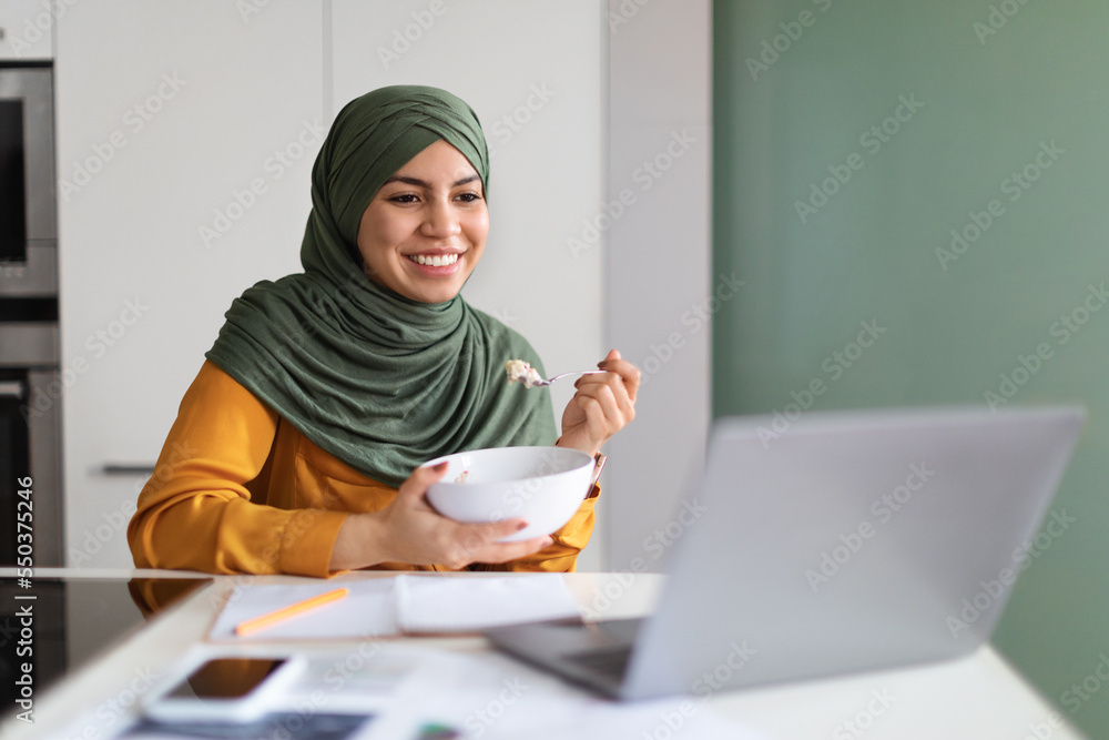 Young Muslim Woman Watching Videos On Laptop While Eating Breakfast In Kitchen, Young Muslim Woman Watching Videos On Laptop While Eating Breakfast In Kitchen,