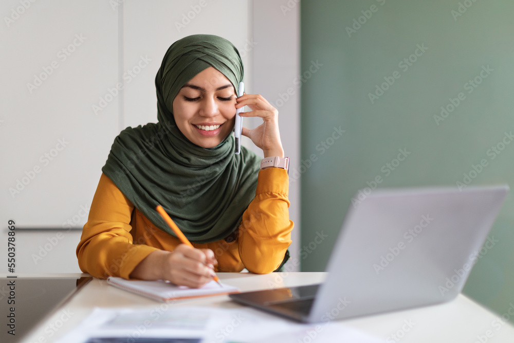Smiling Muslim Woman In Hijab Talking On Cellphone And Noting Down Information Smiling Muslim Woman In Hijab Talking On Cellphone And Noting Down Information
