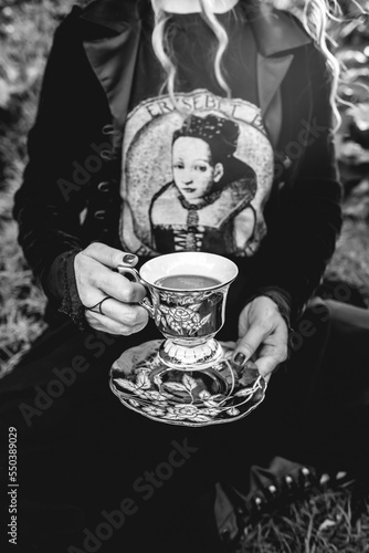 Hands of a girl with dark clothes (and no head) holding a old centuries tea cup and saucer in the park (in black and white)