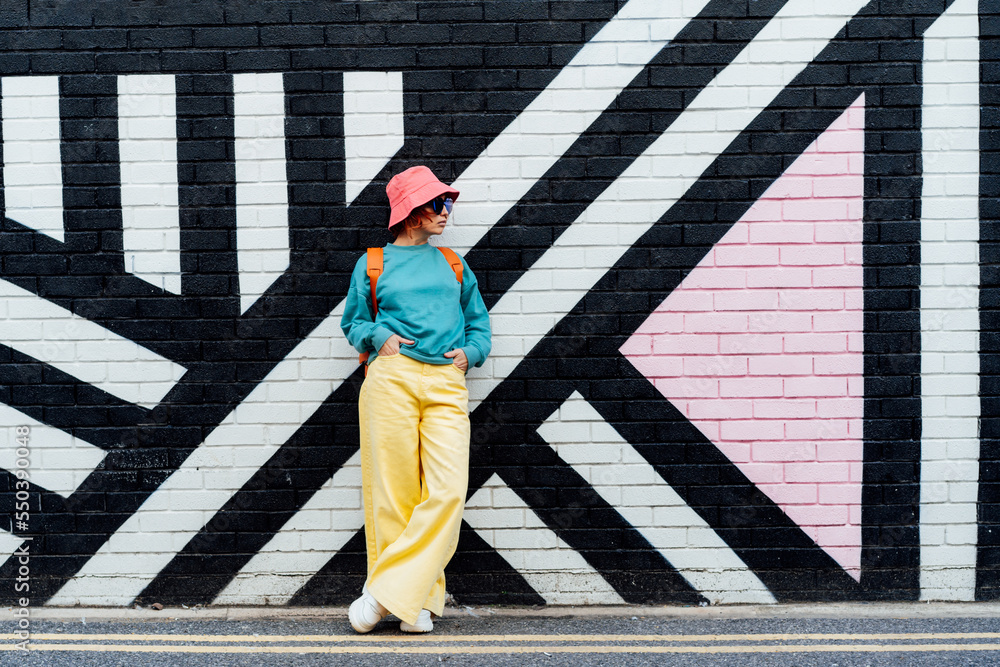 Hipster fashion young woman in bright clothes, sun glasses and bucket hat  posing on the painted brick wall background. Urban city street fashion.  Selective focus. Copy space Stock Photo