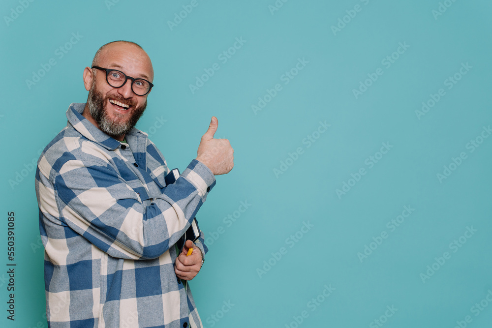Excited caucasian mid adult beardy male in plaid shirt and glasses gestures thumb up toothy smiles against turquoise studio backdrop looks at camera. Mockup, sale, discount, promo. Teacher with book.