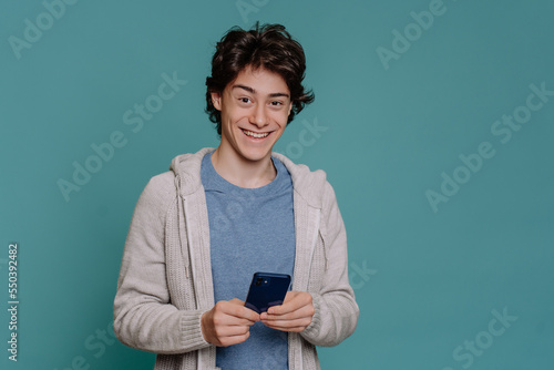 Excited schoolboy in light hoodie and grey t-shirt holds phone looks at camera toothy smiling against turquoise studio background. Brunette guy happy with new smartphone. Mockup, youth, comunications. photo