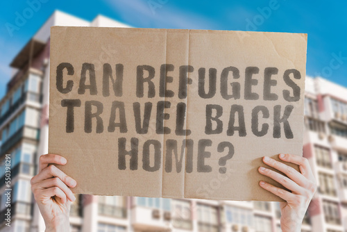 The question " Can refugees travel back home? " is on a banner in men's hands with blurred background. Freedom. War. Ethnic. Government. Human. Liberty. Right. Immigrant. Migration. Regime. Arrest